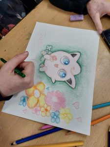 A person is drawing a colorful picture of a cute, cartoon-style cat with big blue eyes, surrounded by pink and yellow flowers and small hearts. The cat is lying on a green background, and the artist's hand is holding a green colored pencil, adding details to the drawing. The workspace includes various colored pencils scattered around on a wooden table. Alt text: A person is drawing a colorful picture of a cute, cartoon-style cat with big blue eyes, surrounded by pink and yellow flowers and small hearts. The cat is lying on a green background, and the artist's hand is holding a green colored pencil, adding details to the drawing. The workspace includes various colored pencils scattered around on a wooden table. CopyS