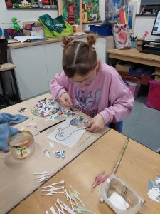 A young girl with glasses and hair styled in two buns, wearing a pink sweatshirt with a cartoon character, is working on an art project at a cluttered table. She is carefully gluing shiny, iridescent paper pieces onto a drawing of a whale on a white sheet of paper. The table is scattered with torn paper, scissors, a paintbrush, and a small container of glue. The background shows an art classroom with colorful artworks, supplies, and storage shelves