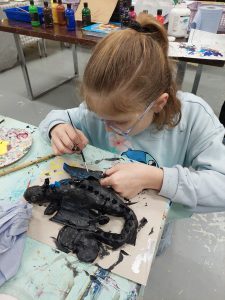 A young girl with red hair tied in a ponytail, wearing safety glasses and a light blue sweatshirt, is working on painting a black crocodile sculpture. The sculpture appears to be made of a flexible material, possibly cardboard or papier-mâché, and is laid out on a table covered with a paint-splattered cloth. The girl is carefully adding details with a small brush, focusing intently on her artwork. In the background, there are art supplies, bottles of paint, and various tools scattered on a table.