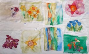  A collection of nine silk paintings of flowers and landscapes, arranged in a grid pattern. The artworks feature various floral designs, including pink, yellow, orange, and purple flowers with green leaves, as well as abstract landscapes with colorful, flowing lines and soft backgrounds. The paintings are on light, textured fabric or paper, creating a delicate and vibrant display of botanical and scenic themes.