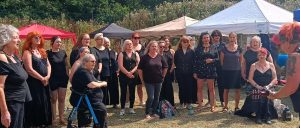 A group singing outdoors during the daytime, standing on a grassy area with trees and bushes in the background. There are several tents in various colors (red, black, beige, white) set up behind them. The women are dressed mostly in black or dark clothing, with some wearing sunglasses, and appear to be performing or participating in a communal event. One woman with red hair and tattoos is playing a guitar on the right side of the image.