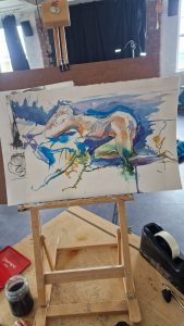 A watercolor painting in progress on a small easel, depicting an abstract, dynamic scene with flowing lines and splashes of blue, green, orange, and purple. The painting appears to be of a reclining figure or animal, with energetic brushstrokes and a sense of movement. The workspace includes a jar of black ink or paint, a red palette, and a tape dispenser on a wooden table. The background reveals an industrial-style studio with brick walls, a window, and studio lighting.