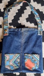 A denim tote bag with colorful patterned fabric accents on the pockets and strap. The bag is laid flat on a black and white patterned surface, with the strap extended. The pockets feature a intricate, multicolored floral and geometric design, adding a decorative touch to the classic blue denim material.