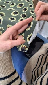  A person is sewing or hemming a piece of fabric with a leopard print pattern, featuring black, tan, and cream spots on an olive green background. The person's hands are visible, holding the fabric and guiding it through the sewing process. They are wearing a gold ring on one finger and a striped sweater with black and beige stripes. The workspace includes a blue surface and a black object nearby.