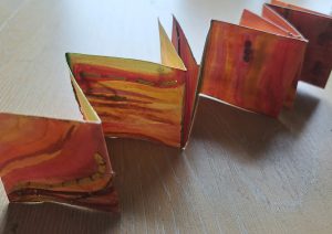 A colorful, handmade accordion-style book or paper craft featuring vibrant, abstract watercolor patterns in shades of orange, red, yellow, and green. The book is partially open, revealing its folded pages, and is resting on a light wooden surface.