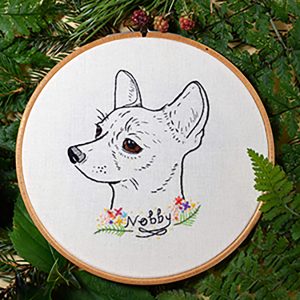An embroidered circular artwork featuring a detailed, realistic drawing of a dog's head, with expressive eyes and upright ears. Below the dog, the name "Nobby" is embroidered in black, surrounded by colorful flowers and green foliage. The embroidery is set against a white fabric background, framed by a wooden hoop, and surrounded by green leaves and small berries.