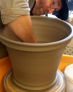 A man with glasses and a cap is working on a large, partially shaped clay pot on a pottery wheel. He is leaning over the pot with his arm inside, shaping or smoothing the interior. The background shows a workshop setting with some tools and materials. A man with glasses and a cap is working on a large, partially shaped clay pot on a pottery wheel. He is leaning over the pot with his arm inside, shaping or smoothing the interior. The background shows a workshop setting with some tools and materials 
