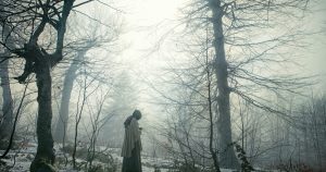 A person wearing a cloak stands alone in a foggy, leafless forest during winter. The scene is mysterious and serene, with tall, bare trees and a soft, diffused light filtering through the fog. 