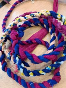 Several colorful fabric braided cords and a fabric strip are arranged on a wooden surface. The cords are in shades of purple, pink, blue, cream, and yellow, with some featuring floral or multicolored patterns. The fabric strip is a solid reddish-pink color. The cords are intertwined and laid out randomly, creating a vibrant, textured scene.