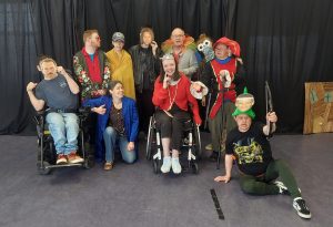 A group of eleven people, some in costumes and some in regular clothing, posing on a stage with a black curtain backdrop. The group includes individuals in wheelchairs, wearing a mix of casual and theatrical outfits, with some wearing masks, wigs, and accessories. They appear to be participating in a theatrical or costume event, with some smiling, gesturing, or making expressive poses