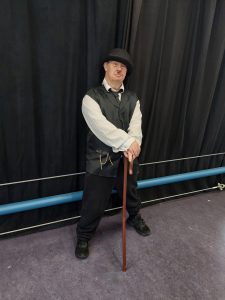 A man dressed in vintage-style clothing, wearing a black bowler hat, white shirt, black vest with a chain, and black trousers. He is standing indoors in front of a black curtain, holding a wooden cane, with a serious expression and arms crossed over the cane