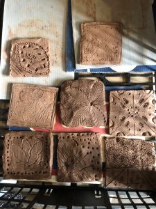This is an image of nine square and one rounded clay tiles with various carved patterns and textures, arranged on a surface with some tools and paper underneath. 