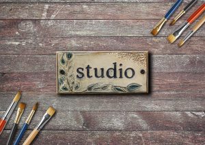 A rectangular ceramic tile with the word 'studio' embossed in the center, decorated with a leafy branch design on the left and bottom edges. The tile is placed on a weathered wooden surface, with several paintbrushes with wooden handles and various colored bristles arranged around it, some at the top right and some at the bottom left