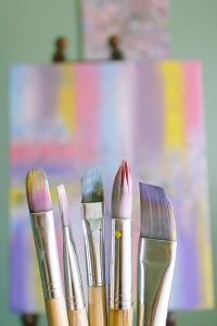A close-up of five paintbrushes with wooden handles and metallic ferrules, standing upright with their bristles painted in various pastel colors. In the background, there is an blurred abstract painting on a canvas, with soft pastel hues and vertical streaks of pink, yellow, blue, and purple, set against a light green wall.