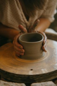 A person shaping a clay pot on a pottery wheel, with their hands covered in wet clay. The focus is on the hands holding the pottery, with the blurred background showing the person's upper body and a patterned shirt