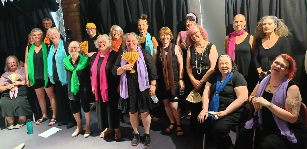 A group of smiling women and a few men, some holding colorful fans, gathered together on a stage with a black curtain backdrop, celebrating at an event or performance. 