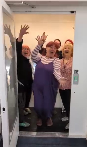 A group of cheerful women, some with red hair and others with blond hair, are happily waving and smiling as they stand together in an elevator doorway, appearing to have fun and enjoying each other's company.




