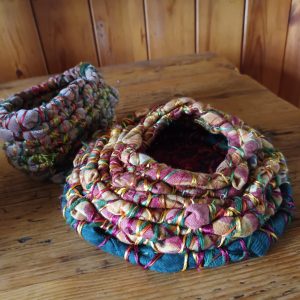 Two colorful, chunky crocheted baskets with open centers sit on a wooden floor in front of a row of wooden cabinets. The larger basket is in the foreground, with a smaller one behind it. 