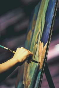 A close-up of a hand holding a stylus and drawing on a large digital display, which shows colorful abstract strokes against a dark background. 