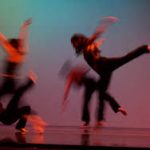 An action photograph of four dancers all dressed in black. They are leaping, the image is motion blurred