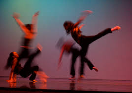 An action photograph of four dancers all dressed in black. They are leaping, the image is motion blurred