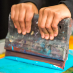 A photograph of 2 hands holding a creen printing for pushing blue ink through a mesh screen