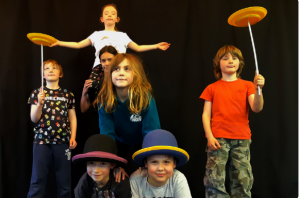 A group of young people in a youth circus workshop. Some children balancing plates. Some children have hats on. Black background