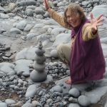 A photograph of Jane Spray with arms out sitting on rocky beach by a small tower of stones