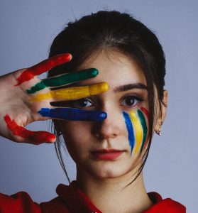 Teenage girl with blue and yellow stripes painted on her face, with coloured paint on her hand draw closely to her face, showing eye through fingers