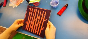 hands holding an orange stencil of tree branches on a brown square board 