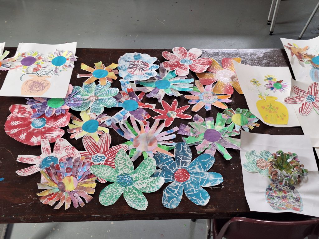 A table covered with colorful, hand-crafted paper flowers and drawings, featuring various patterns and designs. The flowers are made from different types of paper, some with vibrant colors and textured patterns, and are arranged in a somewhat scattered manner. There are also some drawings of flowers on white paper, some with added cut-out elements, creating a lively and creative display.