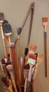 A collection of various paintbrushes with different shapes, sizes, and bristle types, leaning against a light-colored wall. The brushes have wooden handles, some with visible paint stains, and are arranged in a somewhat haphazard manner, creating an artistic and creative atmosphere.
