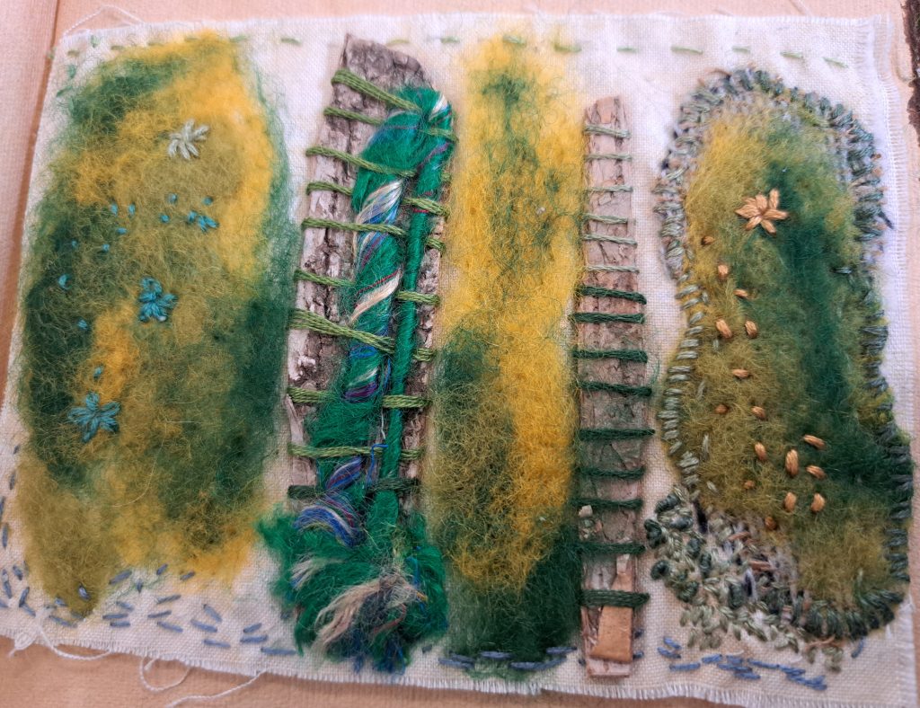 A textile art piece featuring four distinct sections, each with felted wool, embroidery, and natural materials. The sections depict abstract, organic shapes with green, yellow, and blue hues, resembling hills or landscapes. The art is assembled on a piece of fabric with stitched borders, combining various textures and materials for a mixed-media effect.