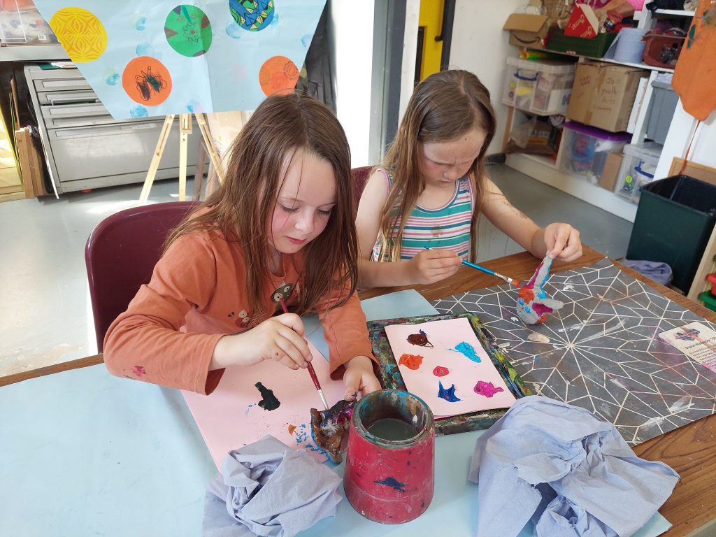 Two young girls are sitting at a table, engaged in painting and decorating a paper mache or clay bunny sculpture. They are using brushes and colorful paints, with palettes of paint in front of them. The background shows a classroom or craft room setting with shelves, storage boxes, and colorful artwork displayed.