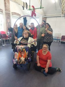 A group of seven people, including a woman in a wheelchair with a colorful blanket featuring a fantasy character, are gathered together in a bright room with high ceilings and exposed brick walls. They are all smiling and posing for the photo, with some standing and others sitting or kneeling. The woman in the wheelchair is holding a hula hoop, and the group appears to be enjoying a fun and lively moment. There are chairs and some hanging ropes or fabric in the background.