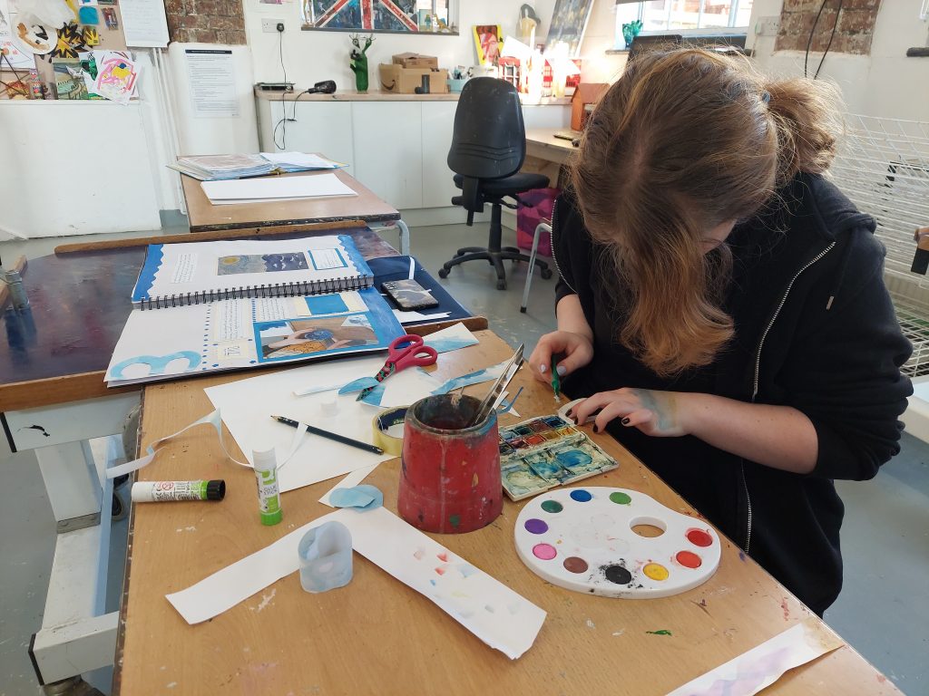  A person with long, wavy hair working on an art project at a cluttered wooden table. They are using a paintbrush and watercolor paints, focusing on their work. The table has various art supplies including a palette with different colors, a jar with brushes, scissors, glue sticks, paper strips, and a cutout paper piece. The background shows an art studio with shelves, artwork, and bright natural light coming through the windows.