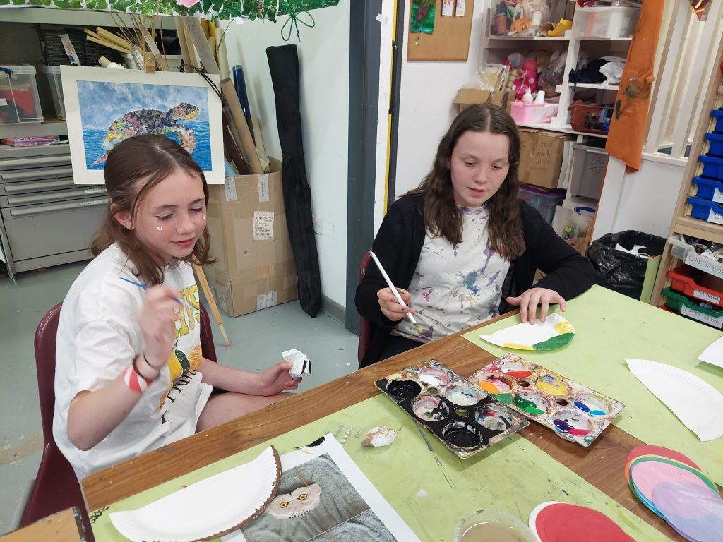 Two young girls sitting at a table in an art classroom, painting paper plates. The girl on the left has light brown hair, is wearing a white t-shirt with colorful graphics, and has small decorative stickers on her face. She is holding a small object in her hand. The girl on the right has darker brown hair, is wearing a black cardigan over a paint-splattered shirt, and is painting a paper plate with green and yellow colors. The table is covered with paint palettes, paper plates, and art supplies, and the background shows shelves filled with art materials and a colorful artwork of a turtle.