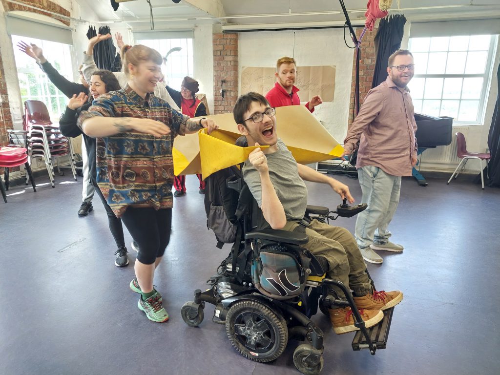A joyful group of people is engaged in a playful activity indoors. The person in the center, seated in a wheelchair, is smiling and holding a yellow paper or fabric object, appearing to enjoy the moment. Surrounding them are others who are smiling, laughing, and participating in the activity, creating a lively and happy atmosphere. The room has large windows, brick walls, and some chairs stacked on the side.