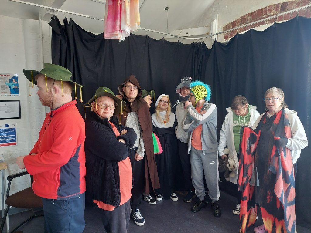 A group of people dressed in various costumes and accessories are standing in front of a black curtain backdrop. Some are wearing hats, including one with a blue wig and yellow striped hat, and others are dressed in cloaks or robes. They appear to be preparing for a performance or participating in a themed event, with a mix of serious and playful expressions.