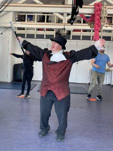 A person dressed in a theatrical costume, wearing a black top hat, a red and black patterned vest, black pants, and white lace cuffs, is standing with arms outstretched in a lively pose. They appear to be performing or acting, with an expressive facial expression. In the background, there are two other people, one on the left in black and one on the right in a blue shirt, in a room with a high ceiling, white walls, and various ropes and equipment hanging from the ceiling.