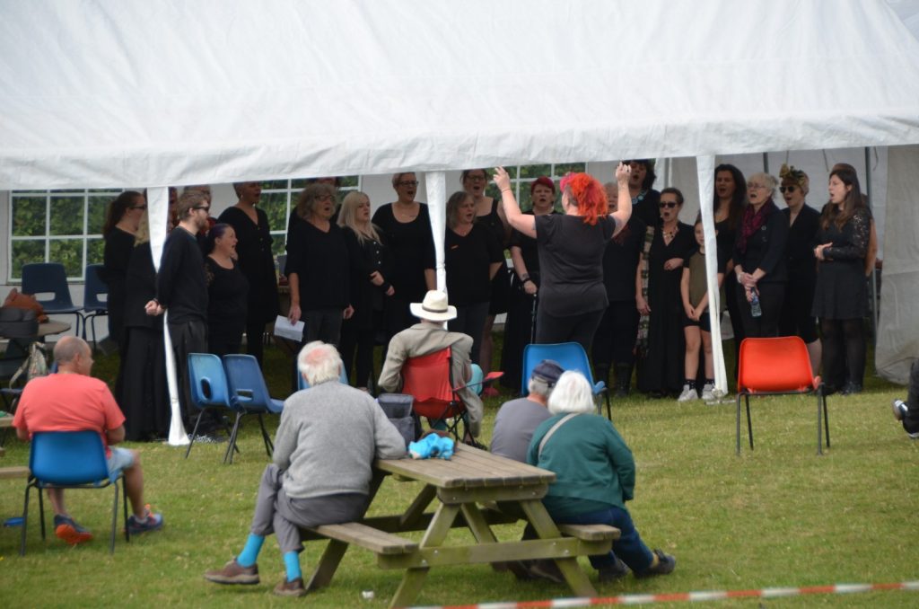 A group of people performing music and singing under a large white event tent outdoors. One person with bright red hair and a guitar is leading the performance, while the choir stands behind them. 