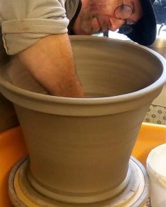 A man is sitting at a pottery wheel, shaping a large, cylindrical clay pot with his arm immersed inside it. The photo is taken from a side angle, focusing on the pot and 