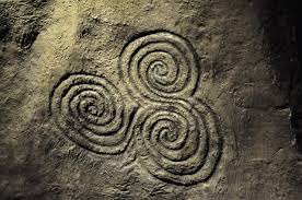 A celtic rock carving featuring three interconnected spiral patterns arranged in a triangular formation. The carvings are etched into a rough, weathered stone surface.