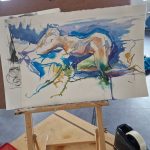 A watercolor painting in progress on a small easel, depicting an abstract, dynamic scene with flowing lines and splashes of blue, green, orange, and purple. The painting appears to be of a reclining figure or animal, with energetic brushstrokes and a sense of movement. The workspace includes a jar of black ink or paint, a red palette, and a tape dispenser on a wooden table. The background reveals an industrial-style studio with brick walls, a window, and studio lighting.