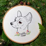 An embroidered circular artwork featuring a detailed, realistic drawing of a dog's head, with expressive eyes and upright ears. Below the dog, the name "Nobby" is embroidered in black, surrounded by colorful flowers and green foliage. The embroidery is set against a white fabric background, framed by a wooden hoop, and surrounded by green leaves and small berries.