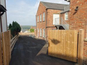 A newly paved, smooth driveway with a wooden fence on the left and a brick building on the right. The driveway extends downhill with a view of houses and trees in the distance under a partly cloudy sky.
