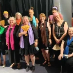 A group of smiling women and a few men, some holding colorful fans, gathered together on a stage with a black curtain backdrop, celebrating at an event or performance.