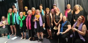 A group of smiling women and a few men, some holding colorful fans, gathered together on a stage with a black curtain backdrop, celebrating at an event or performance.