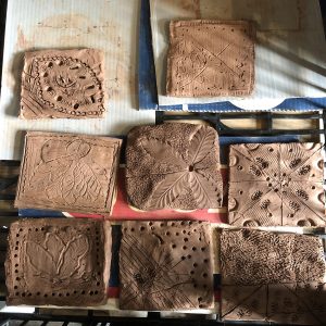 A collection of nine clay tiles with various carved patterns, including leaves, geometric shapes, and abstract designs. The tiles are arranged on a surface, some with detailed textures and others with simple line engravings. The overall appearance suggests they are handmade and in a craft or art workspace.