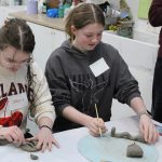 Two young girls are working on clay sculptures at a table in an art classroom. The girl on the left is wearing glasses and a cream-colored sweater with "ISLAND" written on it, and the girl on the right is wearing a gray hoodie with "Molly" on a name tag. They are focused on shaping their clay creations, with various art supplies and materials visible in the background.